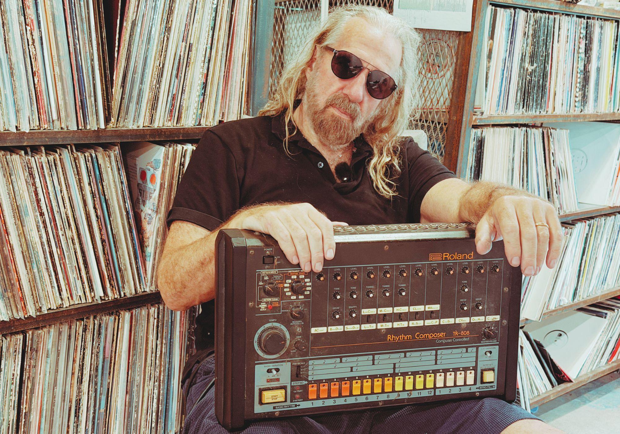 Arthur Baker holding a Roland TR-808 in front of his record collection